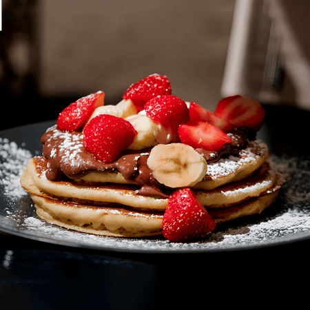 fotografia de pilha de panquecas de banana fit com rodelas de banana e pedaços de morango cobertos por mel e creme de BOLD Whey Milkshake de Baunilha
