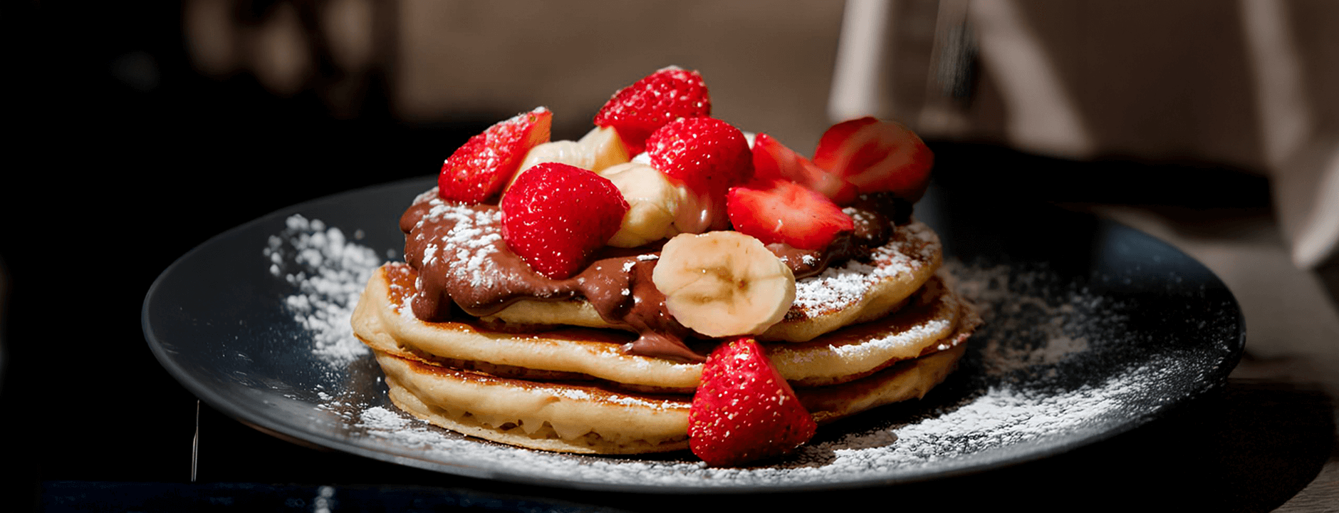 fotografia de pilha de panquecas de banana fit com rodelas de banana e pedaços de morango cobertos por mel e creme de BOLD Whey Milkshake de Baunilha