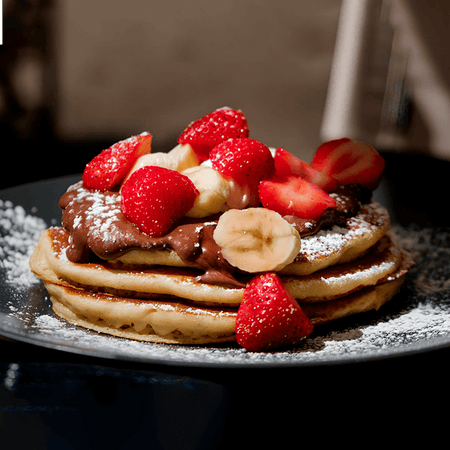 fotografia de pilha de panquecas de banana fit com rodelas de banana e pedaços de morango cobertos por mel e creme de BOLD Whey Milkshake de Baunilha